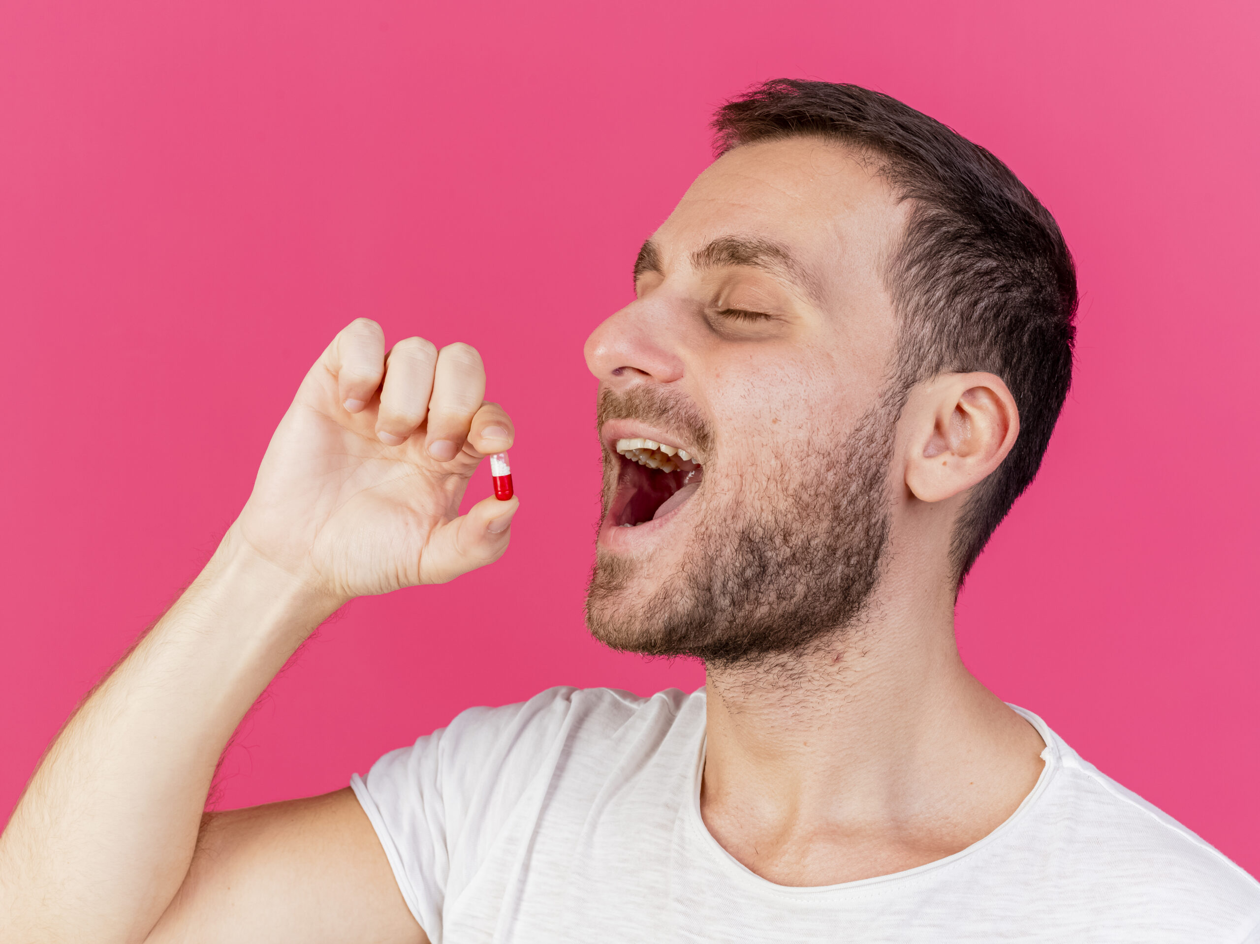 with closed eyes young ill man putting pills in mouth isolated on pink background